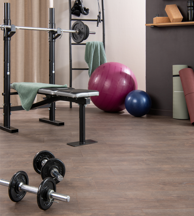 Pièce aménagée en salle de sport à domicile avec banc de musculation, haltères et ballons, sur un sol aspect bois adapté aux contraintes.